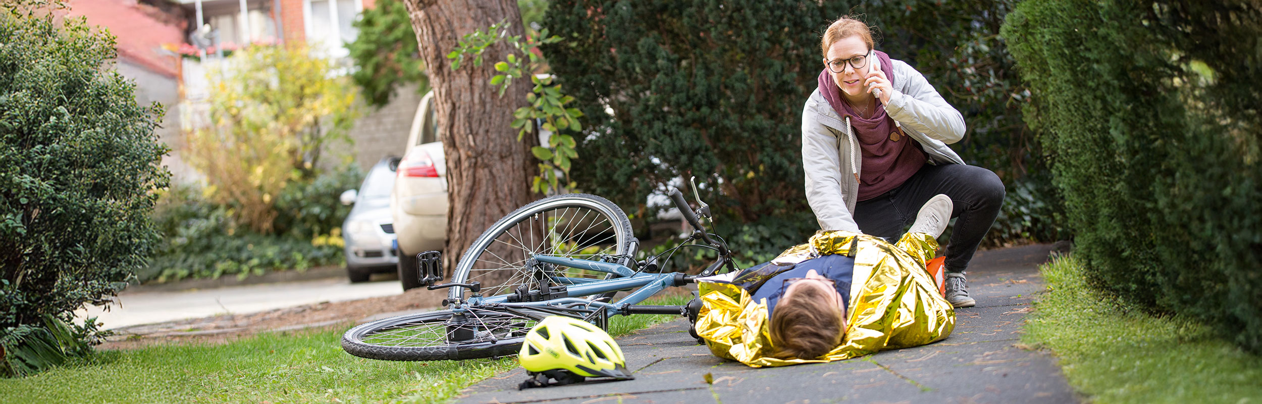 ein verletzter Fahrradfahrer liegt auf einem Weg. Eine Ersthelferin versorgt ihn und ruft telefonisch um Hilfe.